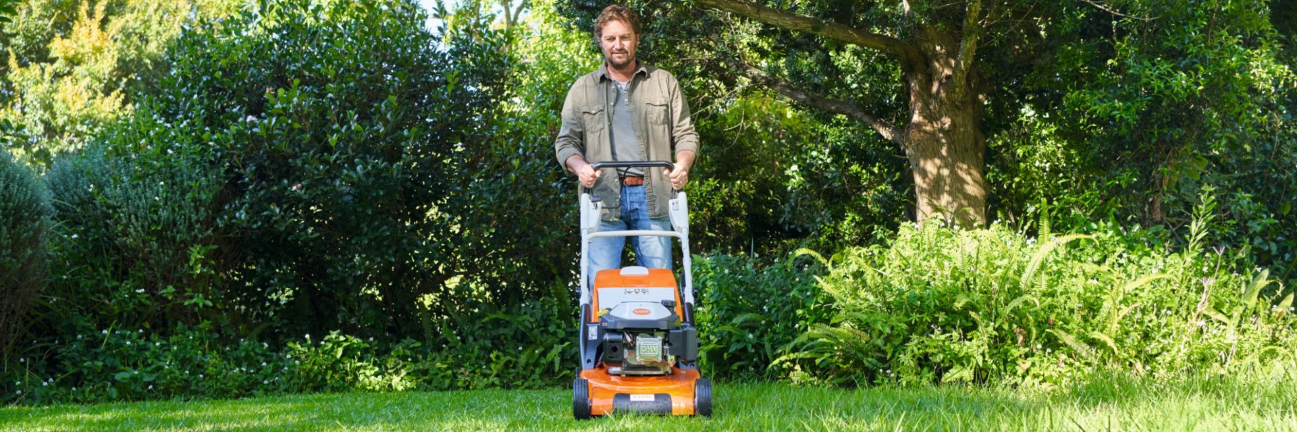 Mann mäht mit elektrischen Rasenmäher von Stihl einen Rasen. Im Hintergrund Bäume und Büsche.